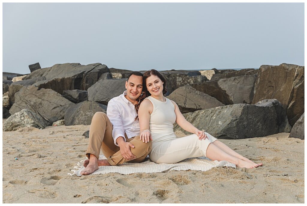 an engaged couple sitting on beach by rock jetty in Asbury Park, Nj